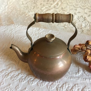 Könnte beinhalten: Eine kupferfarbene Teekanne mit Holzgriff und Deckelknopf. Die Teekanne hat einen gebogenen Ausguss und einen abgerundeten Körper. Der Griff besteht aus hellem Holz und ist mit Metallbügeln am Topf befestigt.