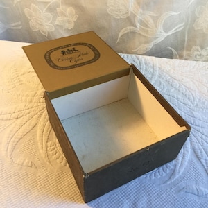 May include: An open, rectangular cigar box with a hinged lid. The box is brown with a white interior. The lid features the text "Thompson Cigar Company Custom Made Cigars" in a circular design.