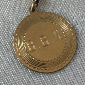 Puede incluir: Un colgante circular dorado con las letras "B.B.X" en el centro. El colgante tiene un borde decorativo con un borde festoneado y está sujeto a una cadena.