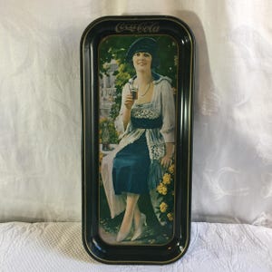 May include: Vintage Coca-Cola advertising tray with a black border. The tray features a woman in a blue dress holding a glass of Coca-Cola. The Coca-Cola logo is at the top. The background shows a garden scene.