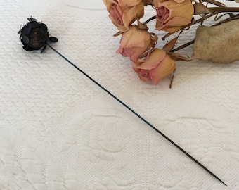 May include: A black metal pin with a round, textured head and a long, thin stem. The pin is lying on a white surface next to a bouquet of dried roses.