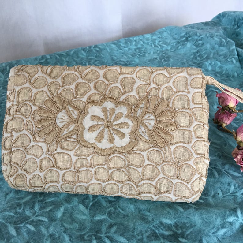 May include: A white clutch purse with a floral design embroidered in a light brown color. The purse has a flap closure and a small handle.