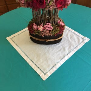 Puede incluir: Arreglo floral con rosas rojas y flores rosas, sobre una base tejida marrón oscuro. El arreglo se encuentra sobre un mantel individual cuadrado de lino blanco con un borde cosido azul, sobre un mantel turquesa.