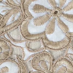 May include: Close-up of a white fabric with beige embroidered floral designs. The fabric has a delicate, intricate pattern.
