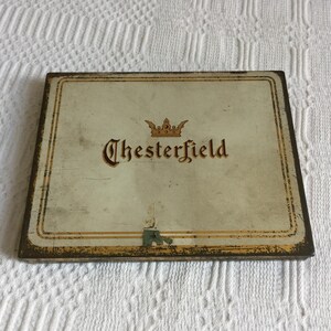 May include: Vintage Chesterfield cigarette tin. The rectangular tin has a cream-colored background with the word "Chesterfield" in brown script, topped with a gold crown. The edges are trimmed with gold and brown.