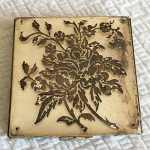 Op de afbeelding: Vierkante compact met een bloemenontwerp. De crèmekleurige achtergrond heeft een gedetailleerd, goudkleurig bloemenpatroon. De randen zijn afgewerkt met een metalen rand. De compact is gesloten en lijkt vintage te zijn.