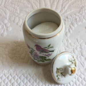 May include: A white ceramic jar with a lid, featuring a gold rim and a painted design of a purple bird and flowers. The jar is open, revealing a white cotton pad inside. The lid is next to the jar.