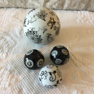 May include: A collection of decorative ceramic spheres in various sizes. The largest sphere is white with black floral patterns. The smaller spheres are black and white, featuring floral and geometric designs. The spheres are arranged on a white, textured surface.