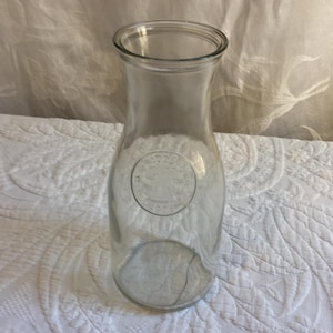 May include: A clear glass milk bottle with a wide base and a narrow neck. The bottle has a circular embossed design on the front. The bottle is empty and sits on a white, textured surface.