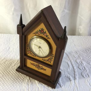 May include: A dark brown wooden mantel clock with a pointed roof and two decorative spires. The clock face is round with a gold-tone bezel, white dial, and black hands. The front panel features a light yellow background with painted floral motifs.