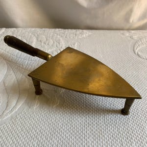 May include: A vintage brass and wood hot iron with a wooden handle and three legs. The iron is shaped like a triangle and has a smooth, polished surface.