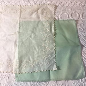 May include: A white square handkerchief featuring intricate white floral embroidery along the scalloped edges, layered over a solid light green square handkerchief with a simple stitched border. Both items are made of thin, lightweight fabric.