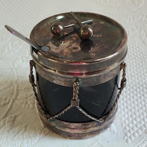 Puede incluir: Un tarro vintage plateado con un cuerpo de vidrio oscuro y una tapa. El tarro tiene forma de tambor con asas decorativas en forma de cuerda y una cuchara pequeña. La tapa presenta dos perillas redondas.
