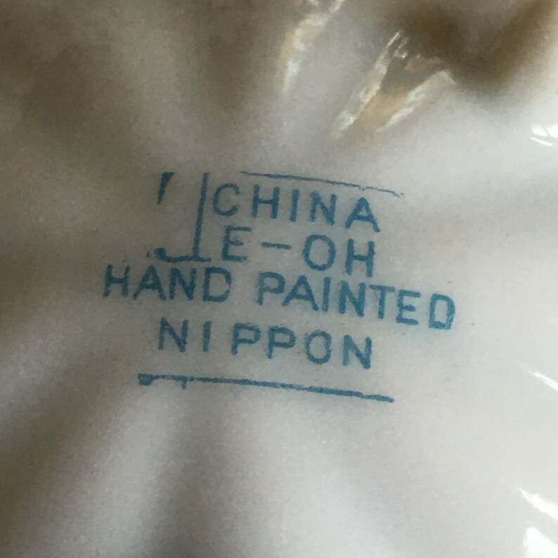 May include: Close-up of a blue stamp on a white surface that reads "China E-OH Hand Painted Nippon".
