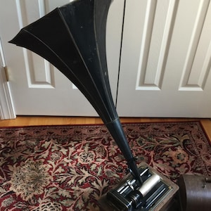May include: An antique gramophone with a large black horn and a wooden base. The horn is angled upwards, and the base features intricate mechanical components. The gramophone sits on a patterned rug, suggesting a vintage aesthetic.