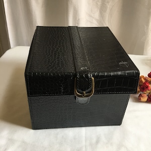 Könnte beinhalten: Eine schwarze Lederbox mit Krokodilprägung und einem goldenen Metallverschluss. Die Box hat einen Klappdeckel und einen dekorativen Riemen.
