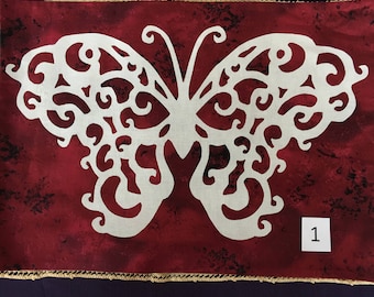 May include: A white, intricate butterfly design is cut out of a red fabric with a dark, speckled pattern. The fabric is bordered with a gold trim. The number "1" is written on a white tag in the lower right corner.