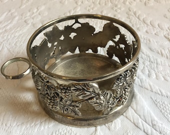 May include: Silver wine bottle holder with a grapevine design. The holder has a handle and a round base.