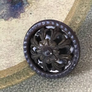 Puede incluir: Un botón redondo de metal plateado con un diseño floral decorativo. El botón tiene un centro elevado y adornado y un borde exterior texturizado.