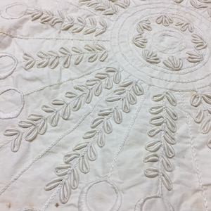 May include: Close-up of a white textile with embroidered leaf and circle patterns. The design features radiating lines and a central circular motif, creating a symmetrical, decorative piece. The fabric appears to be cotton or a similar material.