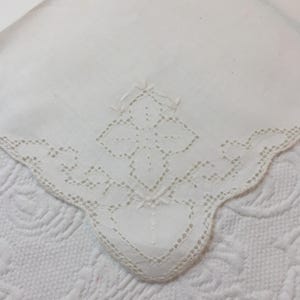 Könnte beinhalten: Weißes Leinentaschentuch mit einem gestickten Blumendesign in der Ecke. Der Rand des Taschentuchs hat eine gewellte, durchbrochene Bordüre. Die Stickerei zeigt ein geometrisches und florales Muster.