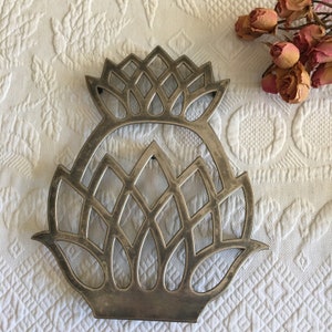 May include: Silver metal trivet in the shape of a stylized pineapple. The trivet has a detailed design with a geometric pattern.