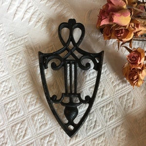 May include: A black cast iron trivet with a decorative design. The trivet is shaped like a shield with a heart-shaped top.