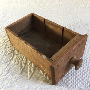 Puede incluir: Pequeño cajón rectangular de madera clara envejecida, con un pomo redondo. La madera muestra vetas visibles y signos de desgaste. El texto "# FINCHING 1714" es visible en el lateral.