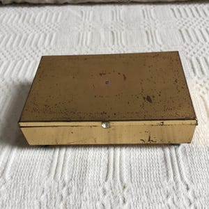 May include: A rectangular, vintage metal box with a hinged lid. The box is painted a faded gold colour with visible signs of wear, including rust spots. A small, round, silver-coloured clasp secures the lid. The box sits on a white, textured surface.