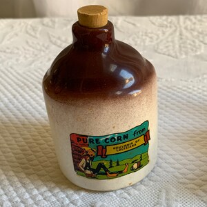 May include: A brown ceramic bottle with a cork stopper. The bottle has a white label with the text "PURE CORN from SOUVENIR of LOUISIANA" and a cartoon image of a man sitting on a log.