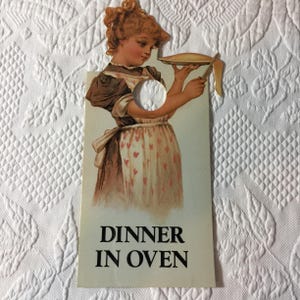 May include: A rectangular door hanger featuring a vintage-style illustration of a person in a brown dress and a white apron with pink hearts, holding a plate with a knife. The text DINNER IN OVEN is printed in black at the bottom of the hanger.