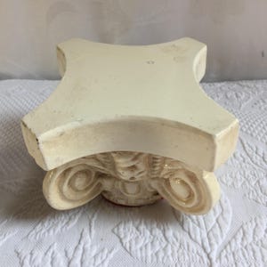 May include: A cream-colored ceramic pedestal stand featuring a square top with concave sides and a base designed with ornate, scrolled volute details. The surface has a smooth finish with subtle variations in tone and visible signs of wear.