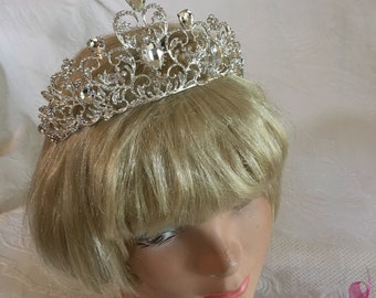 May include: A silver tiara with clear rhinestones and a delicate floral design. The tiara is worn on a blonde wig.