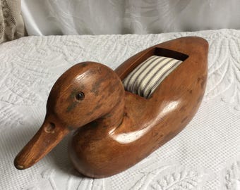 以下が含まれることがあります： 茶色の木製仕上げの、アヒルの形をした針刺し。アヒルは長いクチバシと小さな目を持っています。クッションはストライプの生地で満たされています。アヒルは白い表面に置かれています。
