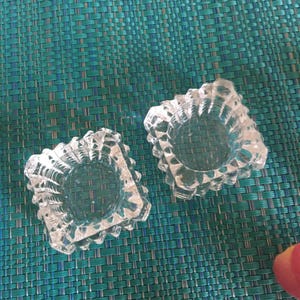 May include: Two clear, square-shaped glass ashtrays with textured, geometric designs. The ashtrays have a central circular indentation and a decorative, jagged edge. They sit on a teal woven surface.