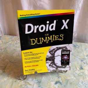 以下が含まれることがあります： 「Droid X for Dummies」というタイトルの黄色と黒の表紙の本で、Droid Xの携帯電話の写真が載っています。本は銀色のスタンドの上に置かれています。本の表紙には、「Making Everything Easier!」と「Learn to:」という文字と、機能のリストが書かれています。