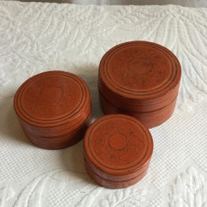 Puede incluir: Tres cajas redondas apiladas de diferentes tamaños. Las cajas son de color marrón rojizo con un diseño decorativo grabado en las tapas. Las cajas están dispuestas sobre una superficie blanca texturizada.