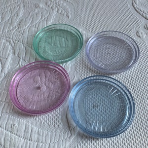 Könnte beinhalten: Vier runde, klare Glasuntersetzer mit einem strukturierten Design. Die Untersetzer sind in den Farben Grün, Lila, Rosa und Blau.