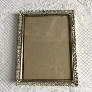 Könnte beinhalten: Ein goldfarbener Bilderrahmen aus Metall mit einem filigranen, komplizierten Design. Der Rahmen ist rechteckig und hat eine leicht abgenutzte Oberfläche.