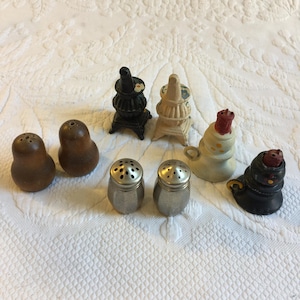 May include: A collection of seven vintage salt and pepper shakers. The shakers are shaped like wood-burning stoves and are made of ceramic and metal. The shakers are in various colors, including black, white, brown, and red.
