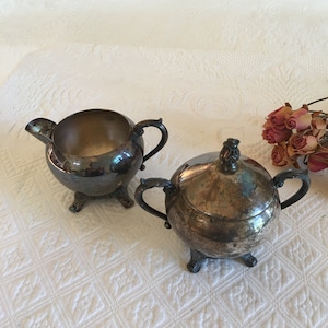 May include: A silver creamer and sugar bowl set with ornate handles and feet. The creamer has a spout and the sugar bowl has a lid.