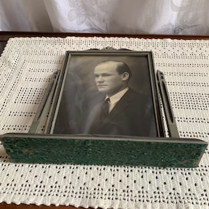 May include: A silver-toned picture frame with a black and white photo of a man wearing a suit and tie. The frame has a green felt backing.