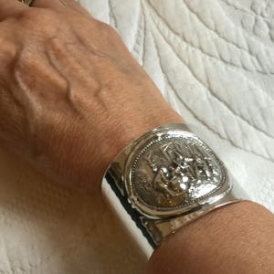 May include: Silver cuff bracelet with a raised design of a group of people. The bracelet is wide and has a smooth, polished finish.