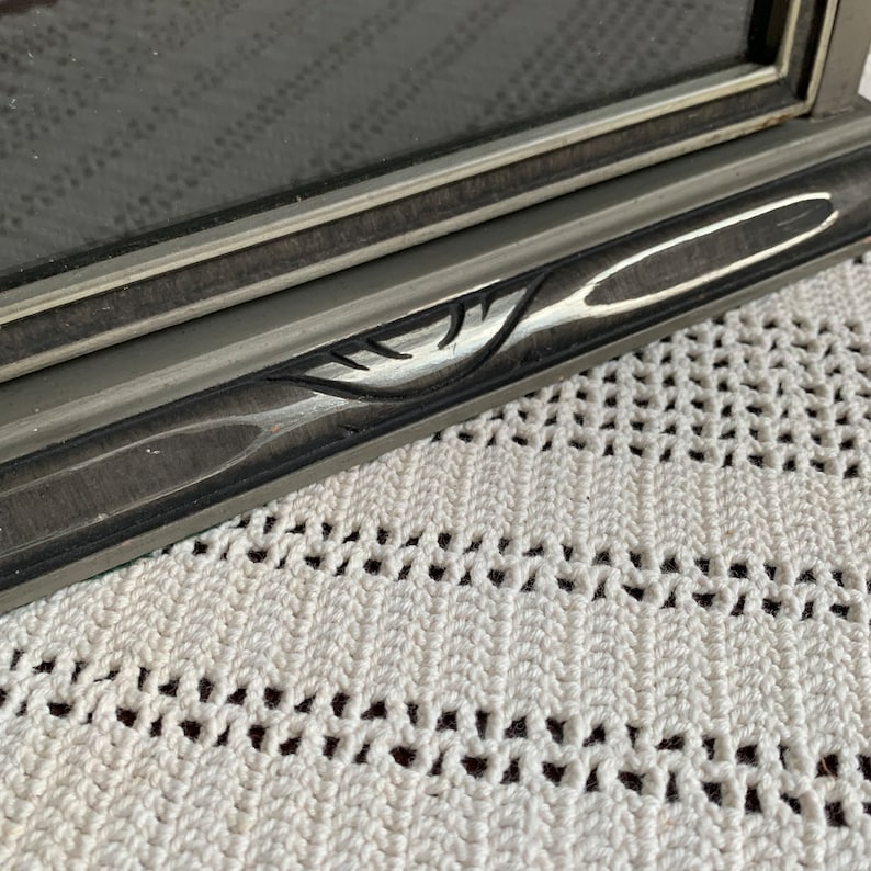 May include: Close-up of a gray picture frame with a silver leaf design on the edge. The frame is resting on a white crocheted surface.