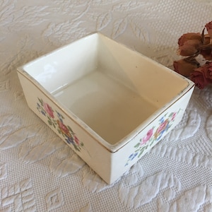 May include: A small, rectangular ceramic trinket box with a white background and a floral design in pink, yellow, blue, and green. The box has a gold trim around the edges.