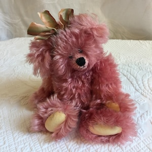 May include: A plush teddy bear with long, shaggy, dusty rose-colored fur. The bear features a small black stitched nose, closed eyes, and light beige fabric pads on its feet. A decorative bow made of iridescent, multi-colored ribbon sits atop its head.