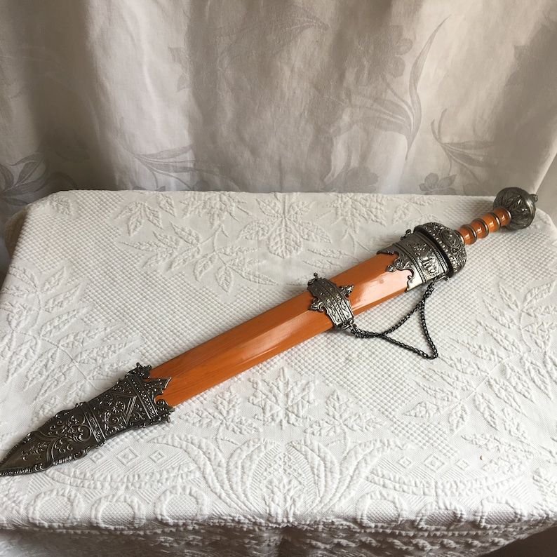 Puede incluir: Una espada decorativa con un mango de madera naranja y detalles de metal plateado. La espada tiene una cadena unida a la vaina.