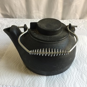 May include: A black cast iron kettle with a round body, a short spout, and a circular lid. The kettle features a silver-colored metal handle with a coiled wire grip section that arches over the top, attached to two small metal loops on the sides.