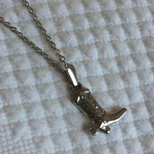 Könnte beinhalten: Ein silberfarbener Metallanhänger in Form eines Cowboystiefels, der an einer passenden dünnen Gliederkette hängt. Der Stiefelanhänger weist geprägte Details entlang des Schafts und einen ausgeprägten Absatz auf, mit einer polierten Oberfläche.