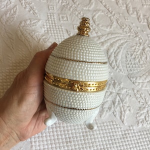 Könnte beinhalten: Weiße Porzellan-Schmuckdose in Eiform mit goldenem Deckel und Füßen. Die Dose ist mit einer perlmuttartigen Textur und goldenen Verzierungen verziert.